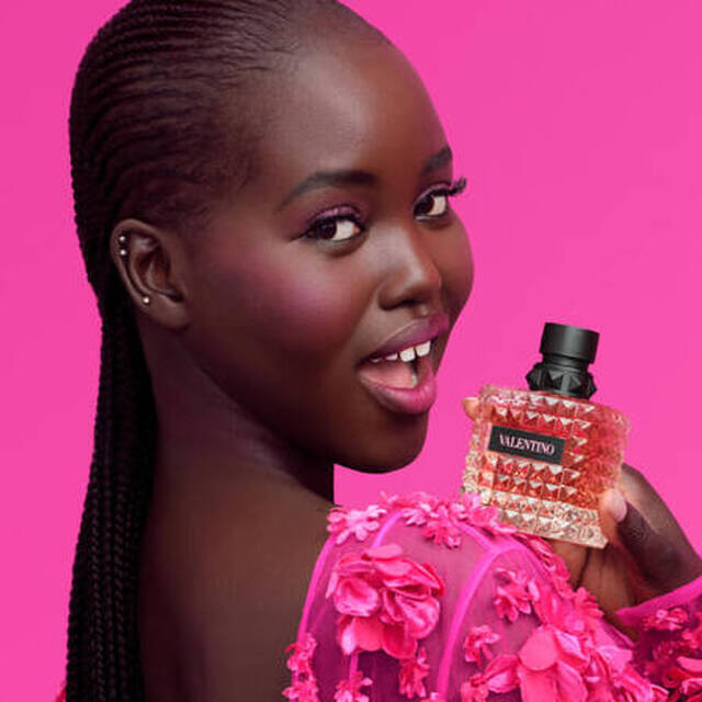 Een vrouw met een stralende lach houdt een parfumfles vast, staande tegen een heldere roze achtergrond. Ze draagt een roze, bloemrijke outfit.