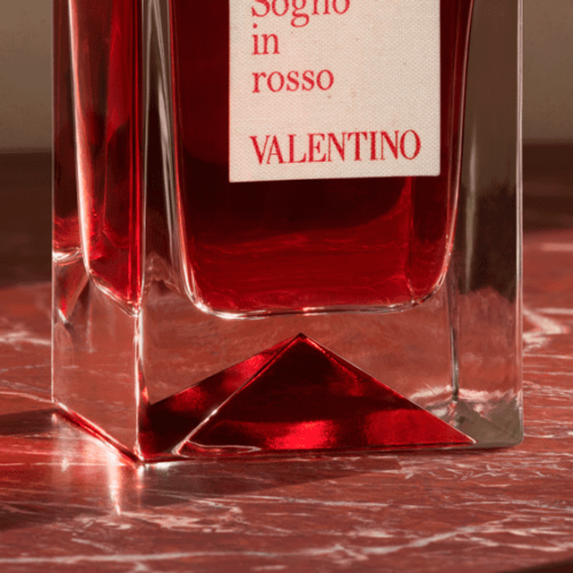 Een close-up van een glas parfumfles met een rode vloeistof en een etiket waarop "Sogno in rosso" en "VALENTINO" staat. De fles staat op een marmeren oppervlakte.
