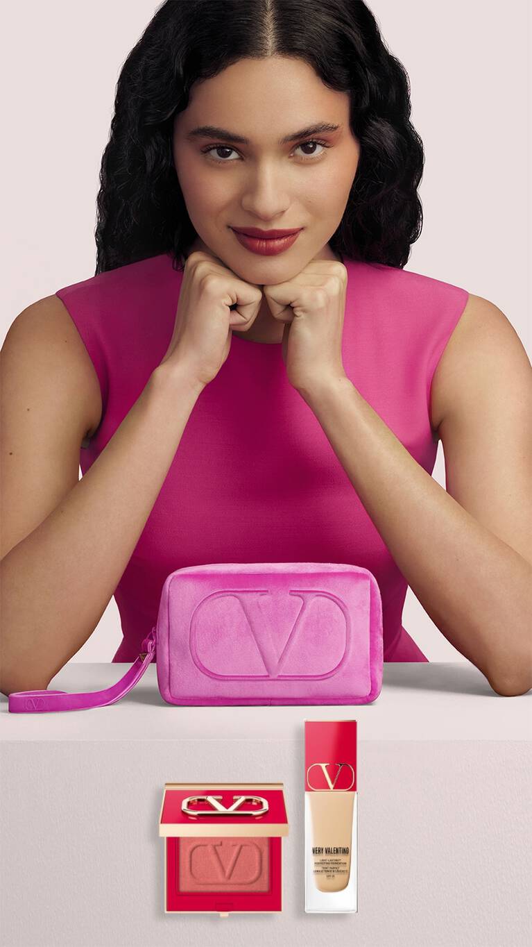 Een vrouw met krullend haar in een fuchsia jurk poseert met make-upproducten en een roze toilettas van Valentino.